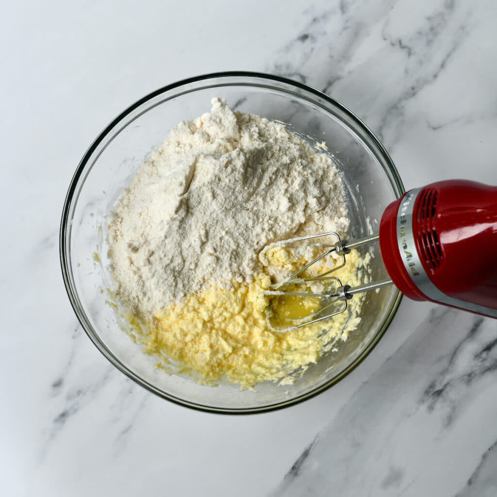 Adding flour to beaten butter, sugar, and egg in a glass bowl.