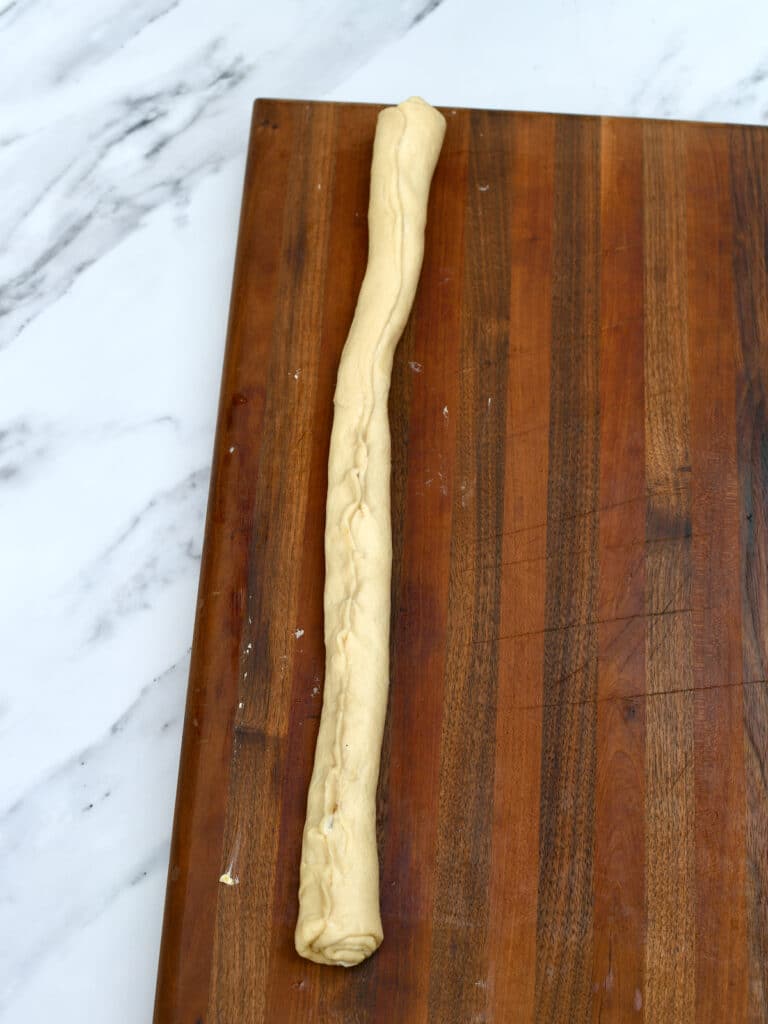 Crescent roll dough rolled into a long log on wooden cutting board.
