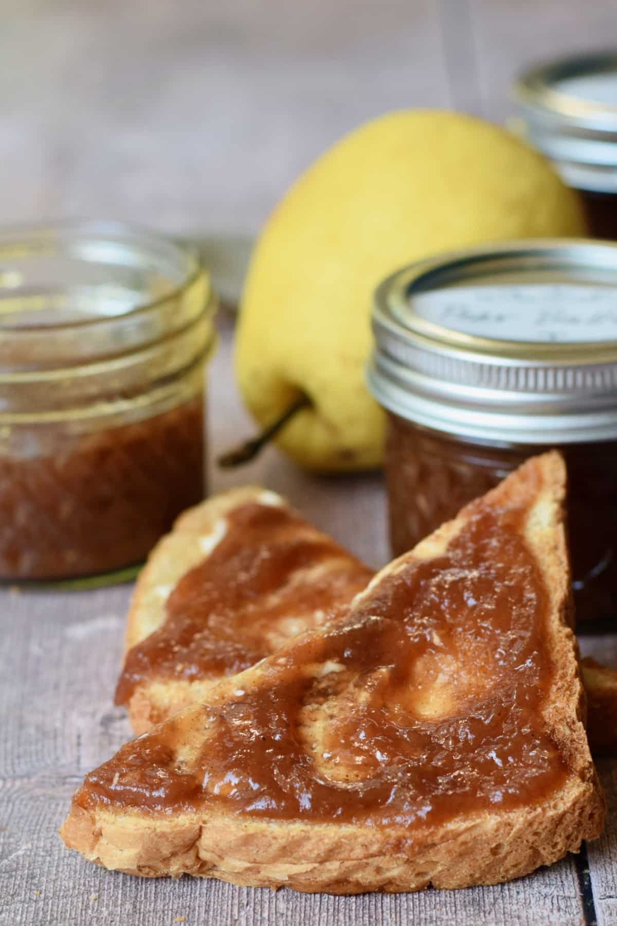 Vertical shot of slow cooker pear butter spread on toast with jars of pear butter in the background.