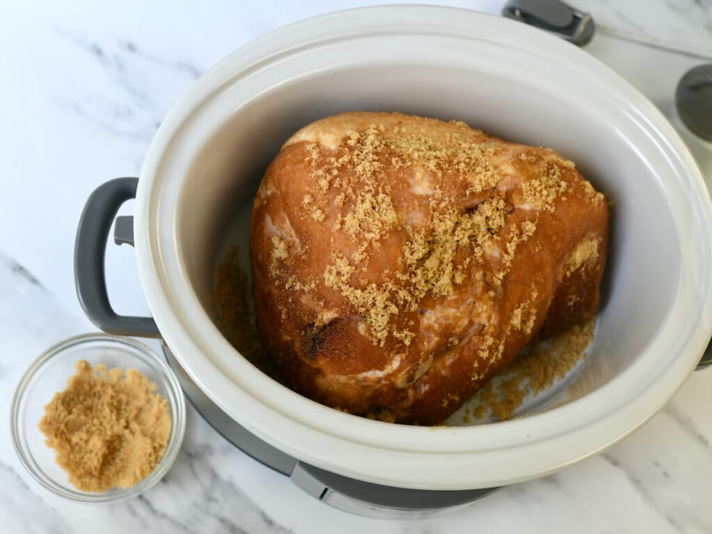 An uncooked ham covered in brown sugar, sitting in the slow cooker.