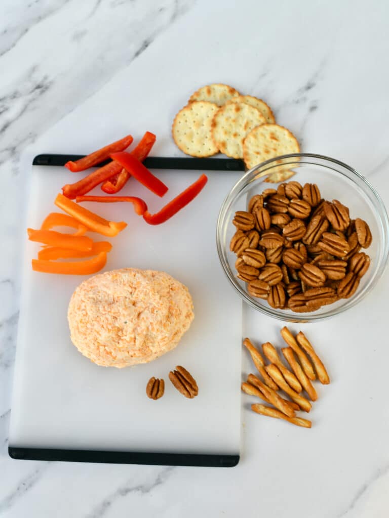 A cheeseball being assembled on a cutting board with pecans, bell pepper strips and crackers.
