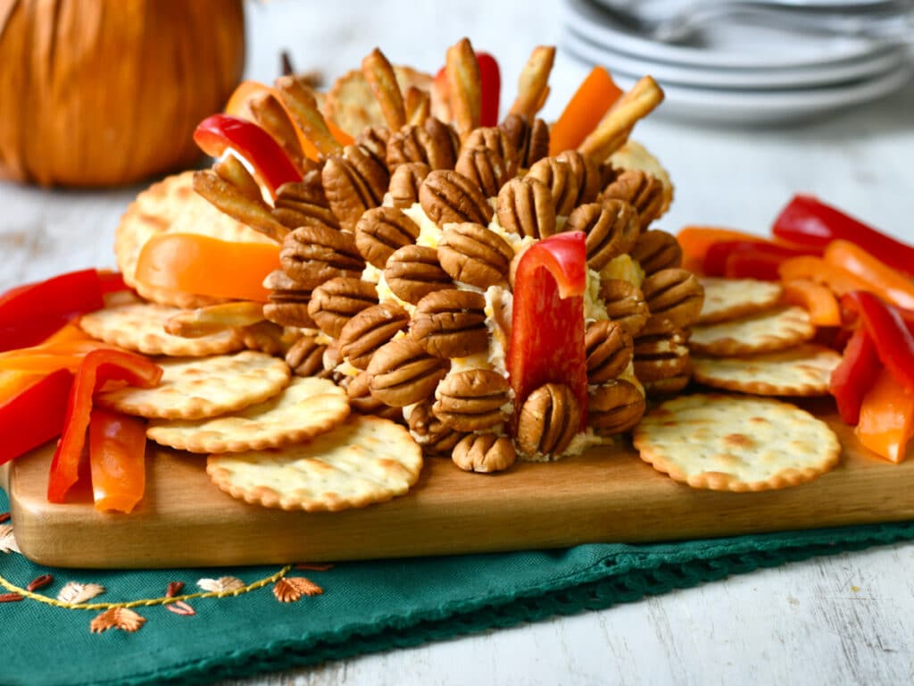 A creative cheese ball designed like a turkey, decorated with pecans and colorful bell peppers for feathers. Surrounded by round crackers on a wooden board with a green cloth underneath. A pumpkin and plates are in the background.
