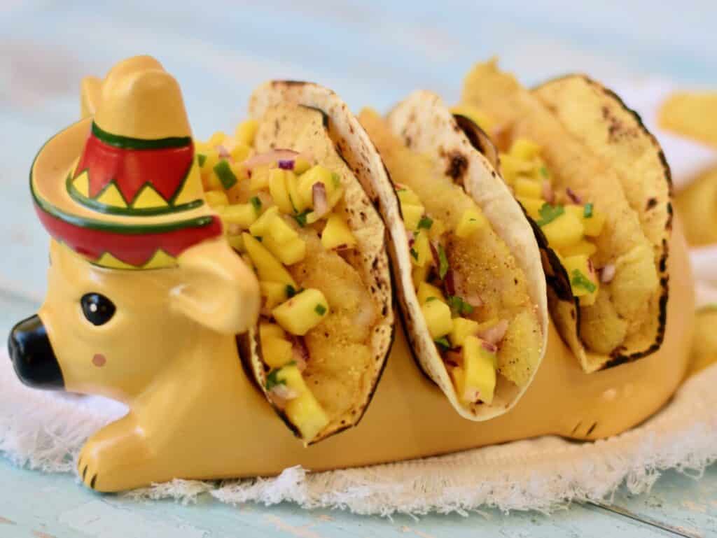 Horizontal image of fish tacos with mango salsa in a taco holder designed like a cow with a sombrero.