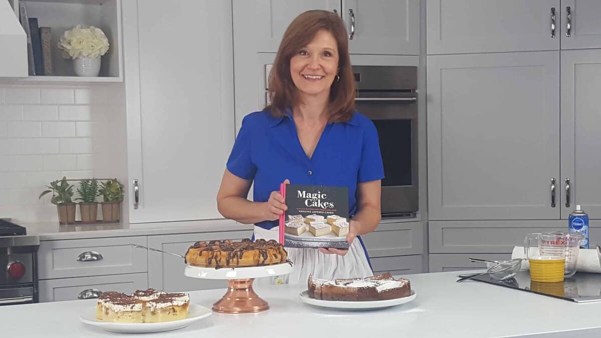Woman in blue shirt holding Magic Cakes cookbook with cakes in front of her.
