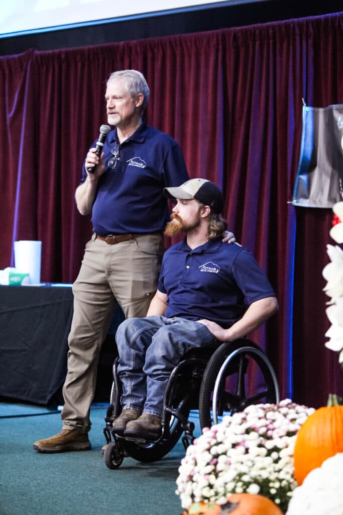 Man on stage with mic from outdoor ability foundation with hand on shoulder of man in wheelchair. scott grayson.