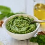 Basil pesto with walnuts in white bowl with spoon and oil in background.