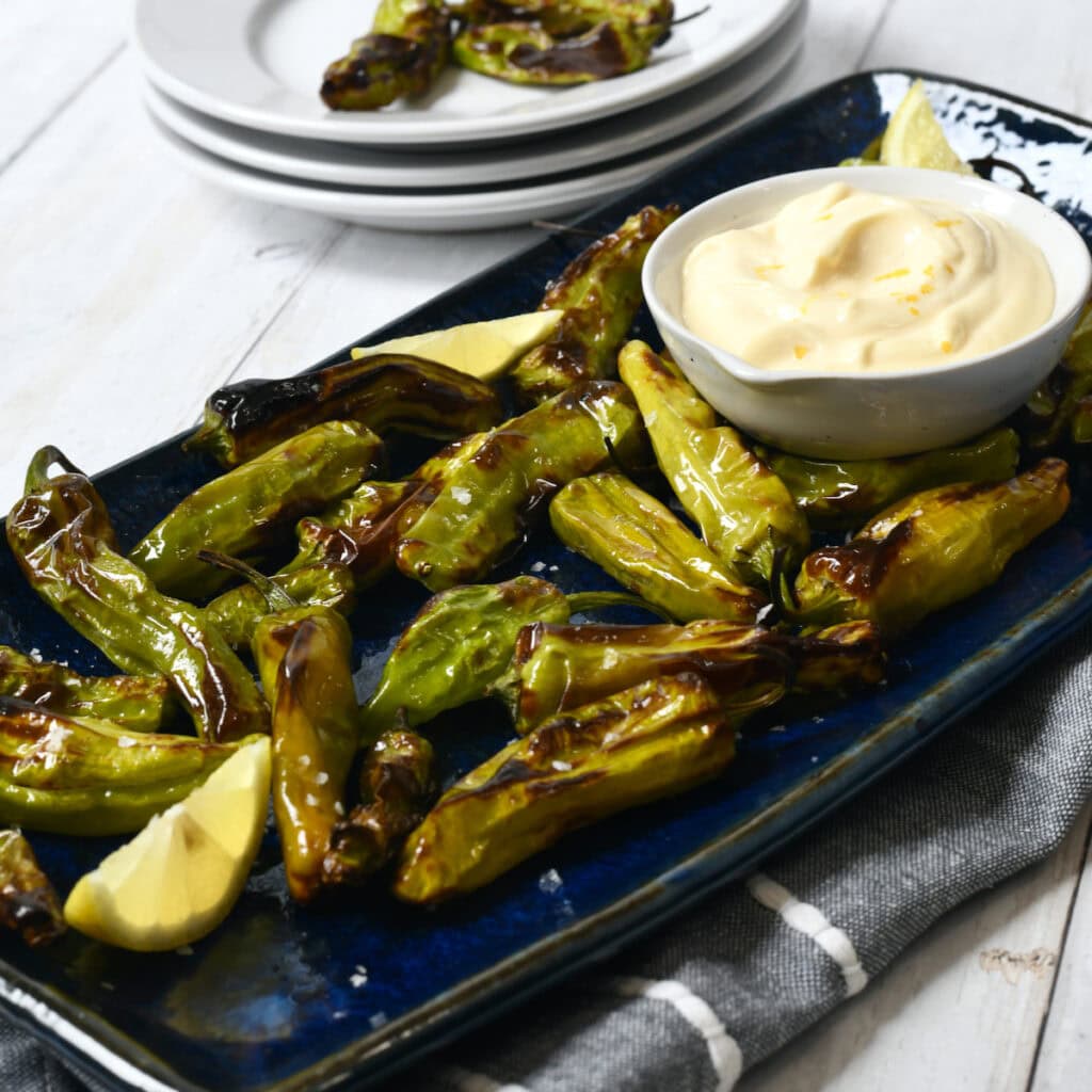 Shishito Peppers roasted in air fryer on blue platter with lemon aioli dipping sauce with stack of white plates in background.