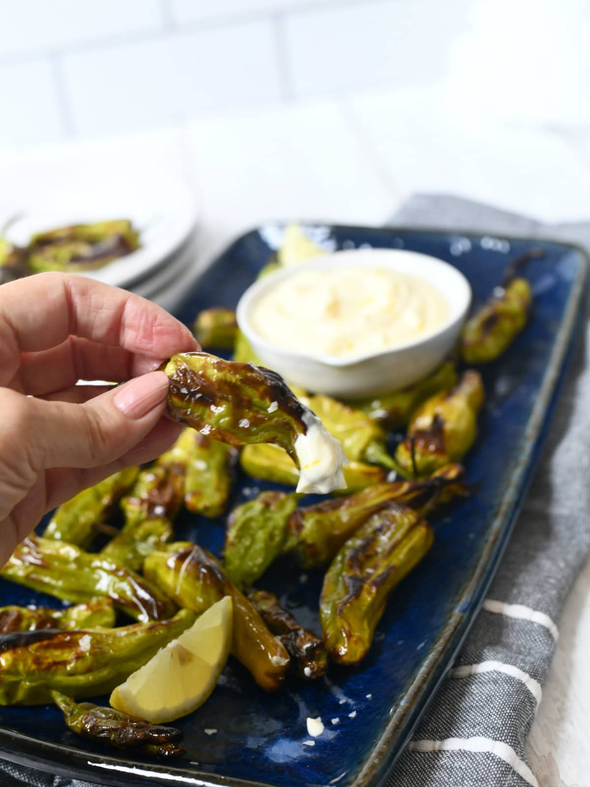 Fingertips holding a roasted Shishito Peppers roasted over a blue platter with lemon aioli dipping sauce.