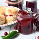 Spoonful of cranberry pepper jelly resting on top of a jar of jelly beside a plate of biscuits and jars of jelly with a jalapeno pepper in the front.