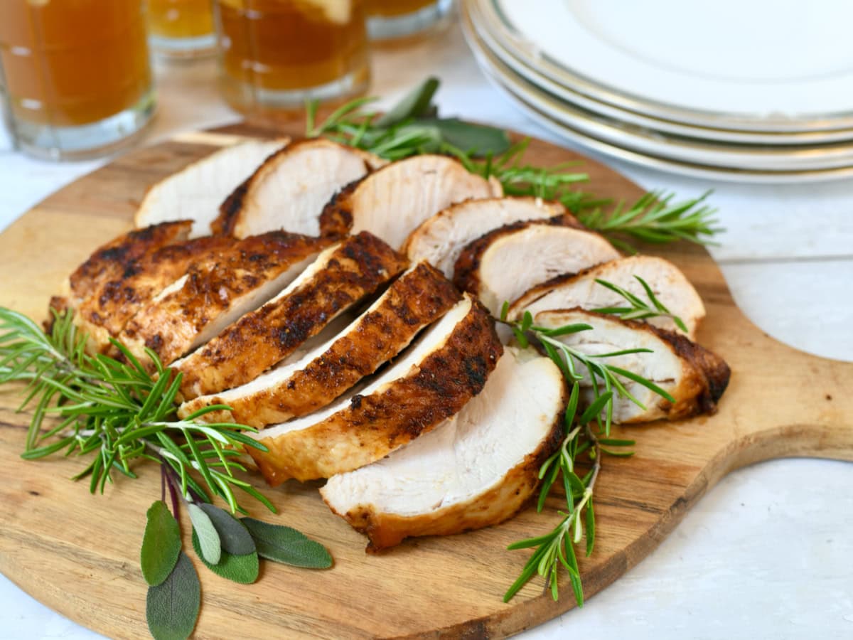 Sliced roasted turkey breast with fresh herbs on wooden cutting board. Tea glasses in background.