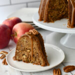Slice of pecan apple bundt cake on a white plate with apples behind it and cake pedestal beside it.