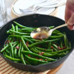 Whole fresh green beans with bacon and brown sugar in a cast iron skillet with spoon dripping sauce on beans.