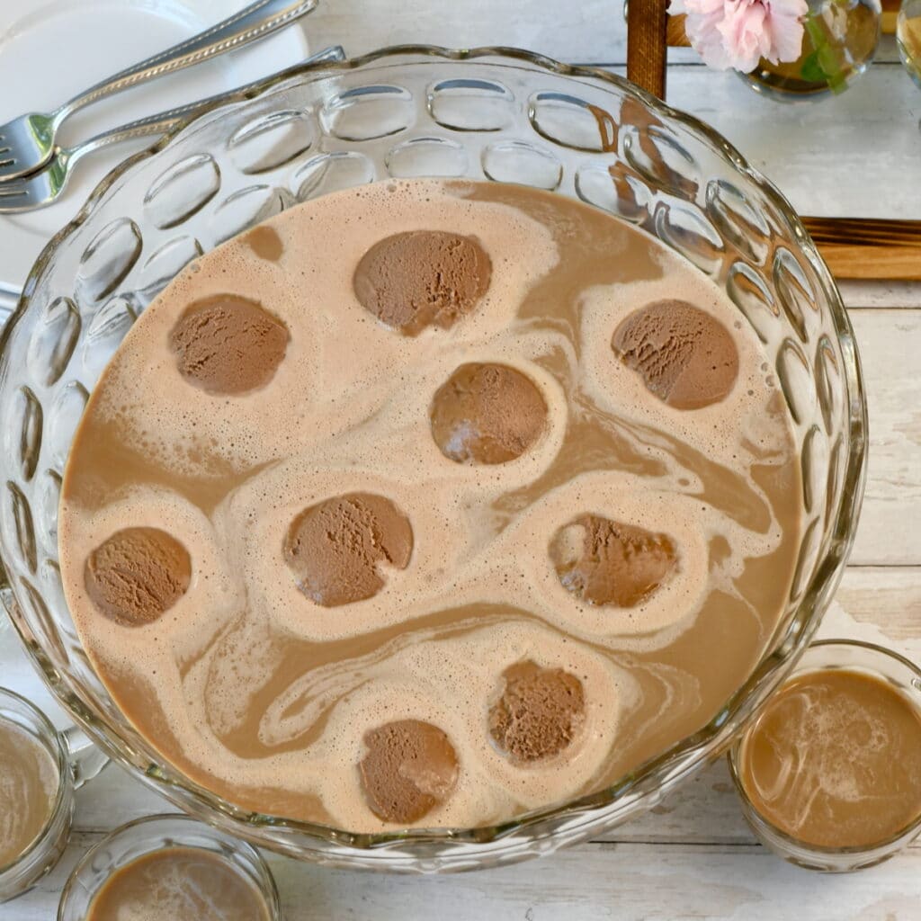 A glass punch bowl filled with creamy coffee punch, topped with scoops of melting ice cream
