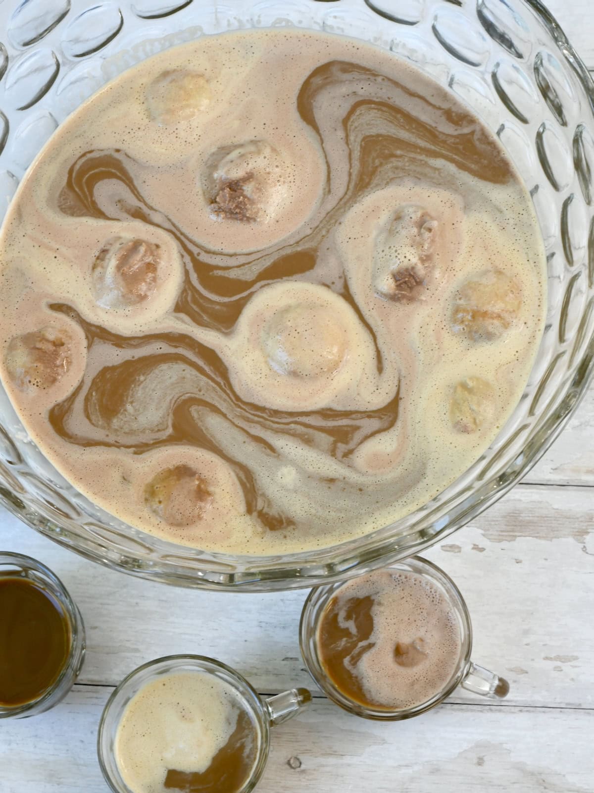 A close-up top view of a glass punch bowl filled with creamy coffee punch, swirled with melting ice cream, beside it are glass cups filled with coffee punch