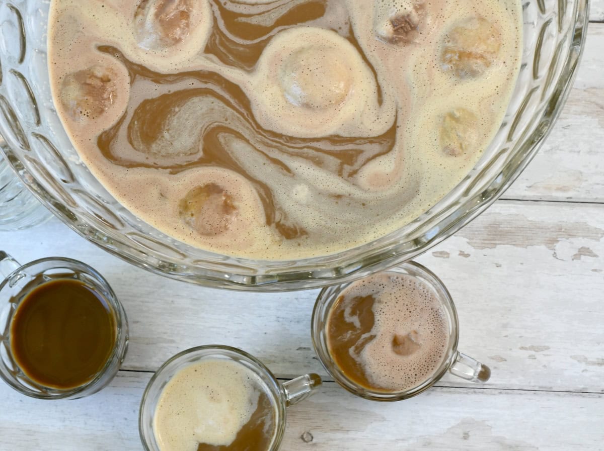 A close-up top view of a glass punch bowl filled with creamy coffee punch, swirled with melting ice cream, beside it are glass cups filled with coffee punch
