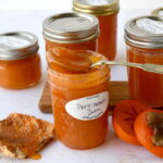 Spoonful of homemade persimmon jam resting on a jar with hachiya persimmon jam spread on toast with a fuyu sliced persimmon next to it.