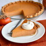 Slice of persimmon pie with pie plate in background and Fuyu persimmon next to it.