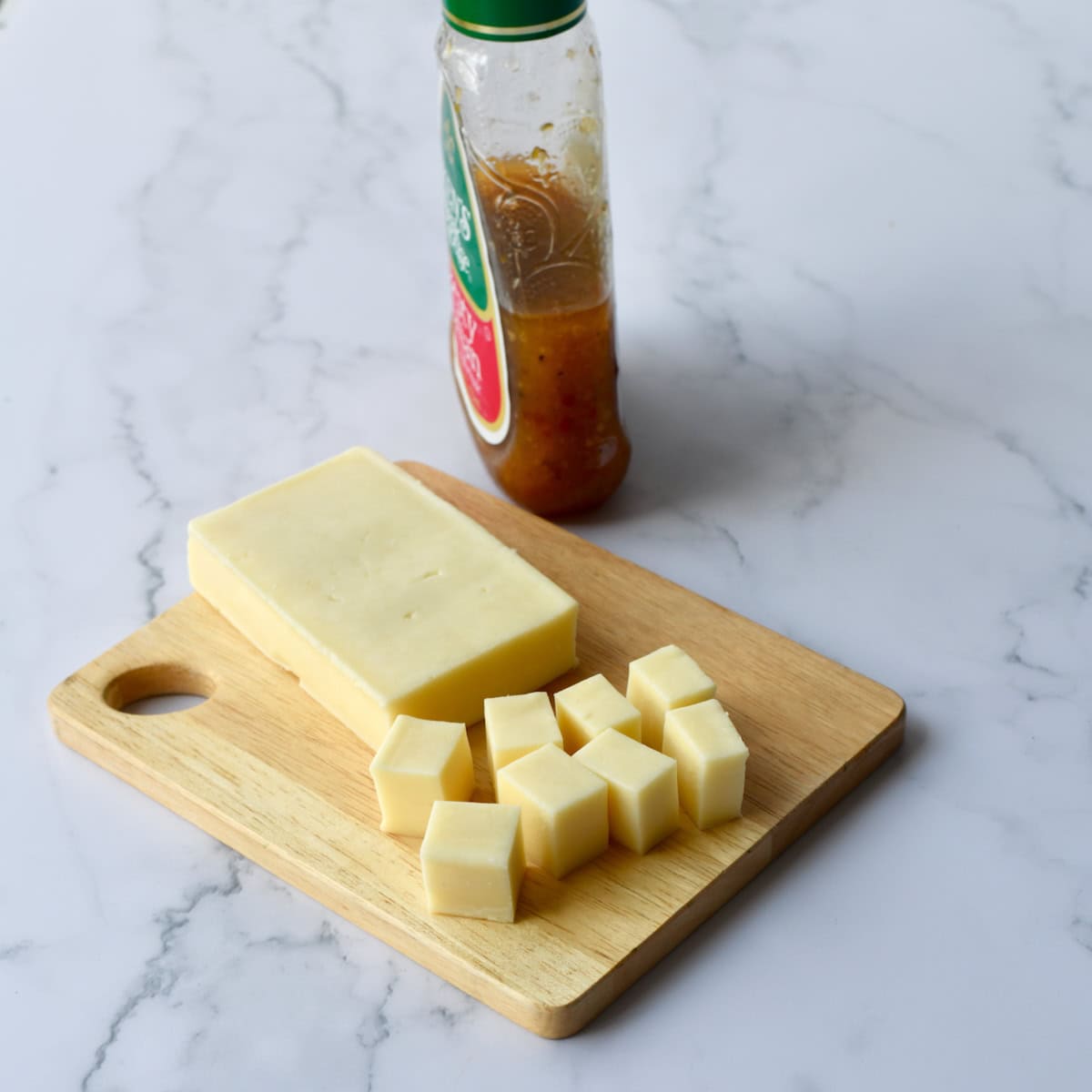 Block of cheese cut into cubes on a wooden cutting board, with a bottle of dressing in the background