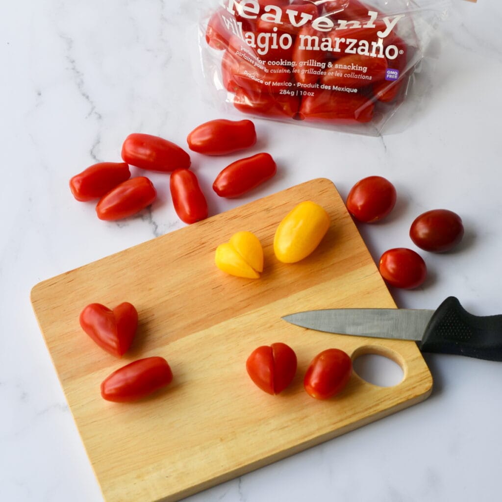Tomatoes, some cut into heart shapes, on a wooden cutting board with a knife and a bag of tomatoes in the background