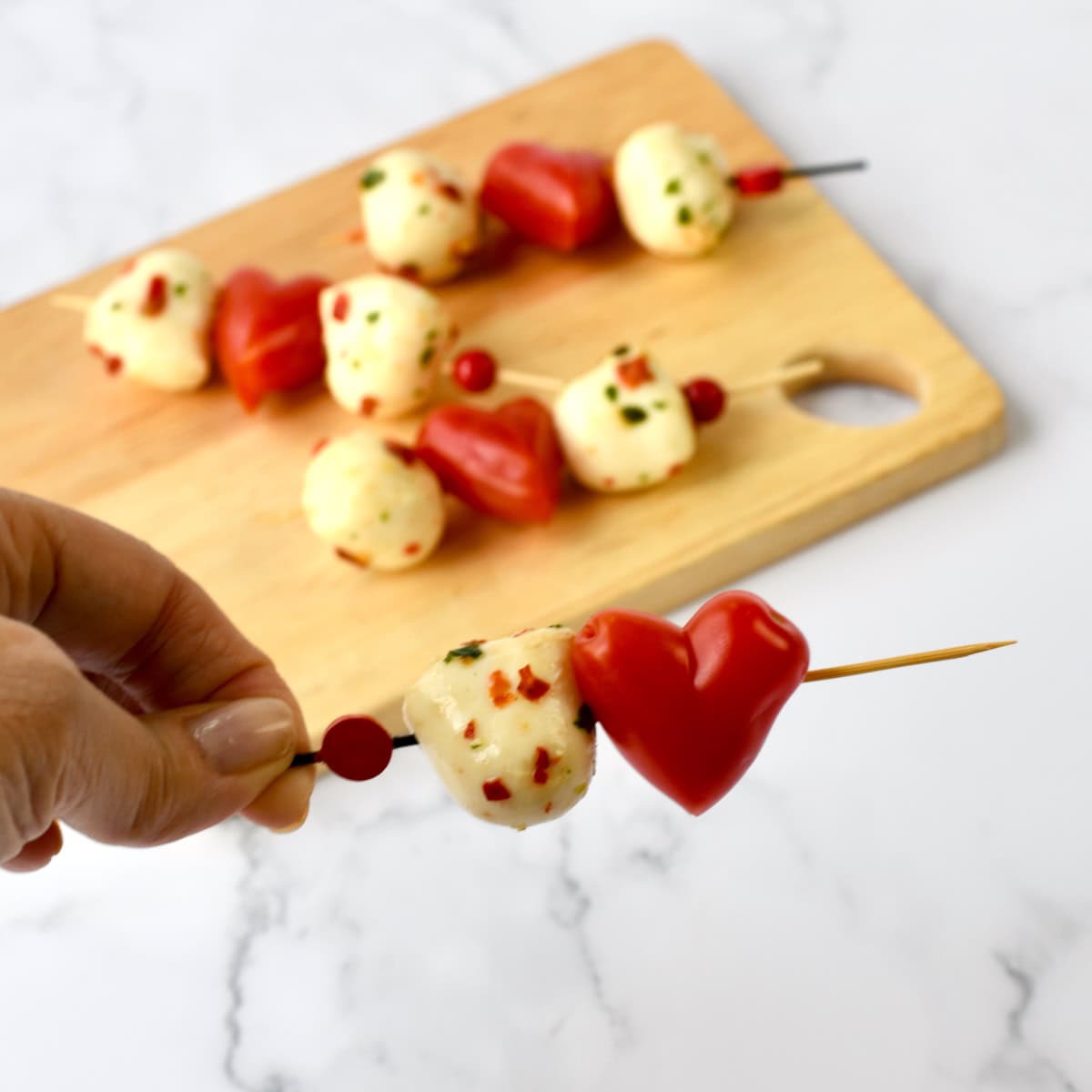 A hand holding a Heart Caprese skewer with mozzarella balls and a heart-shaped tomato, with more skewers on a wooden cutting board in the background