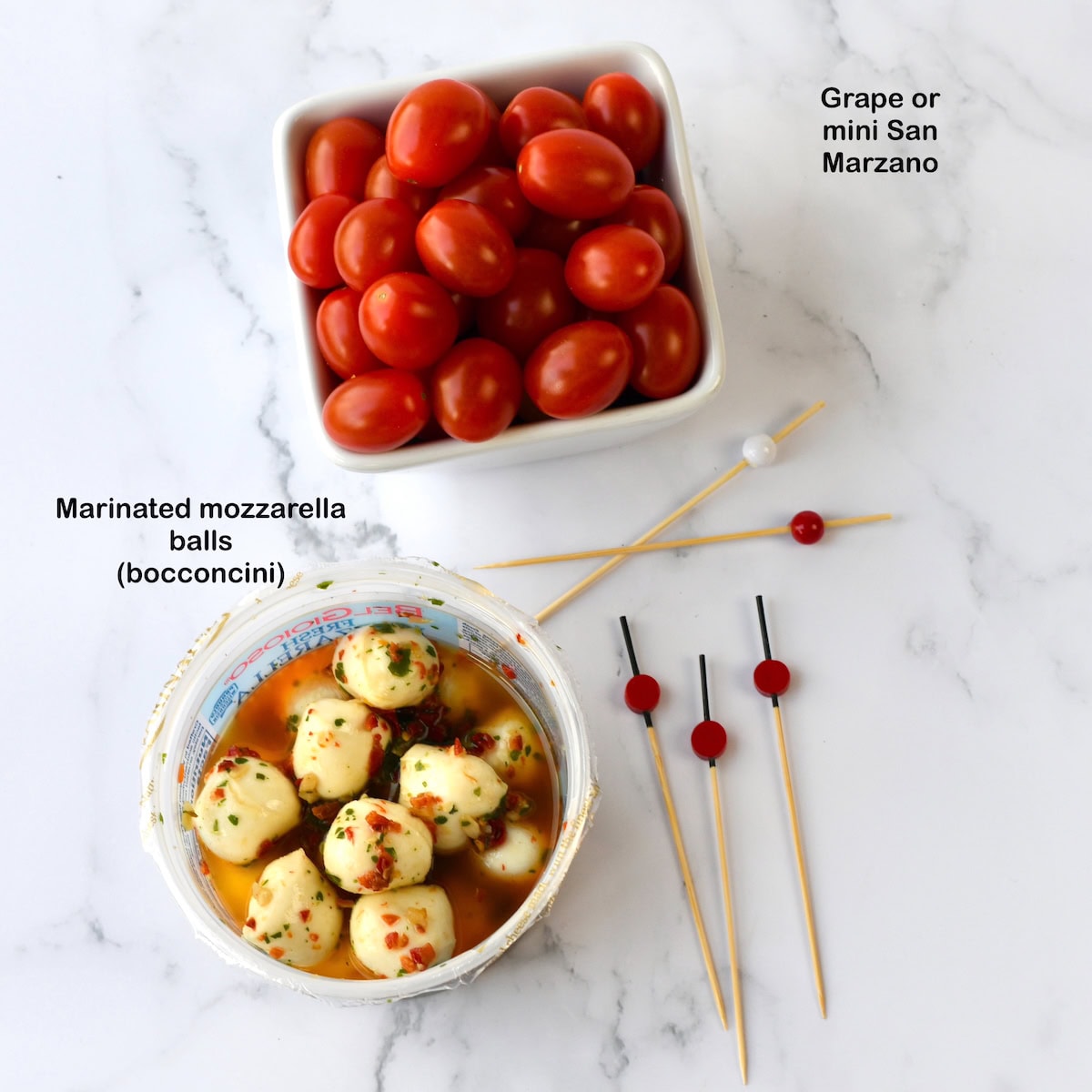 Bowl of marinated mozzarella balls and a dish of tomatoes on a marble surface, with skewers nearby