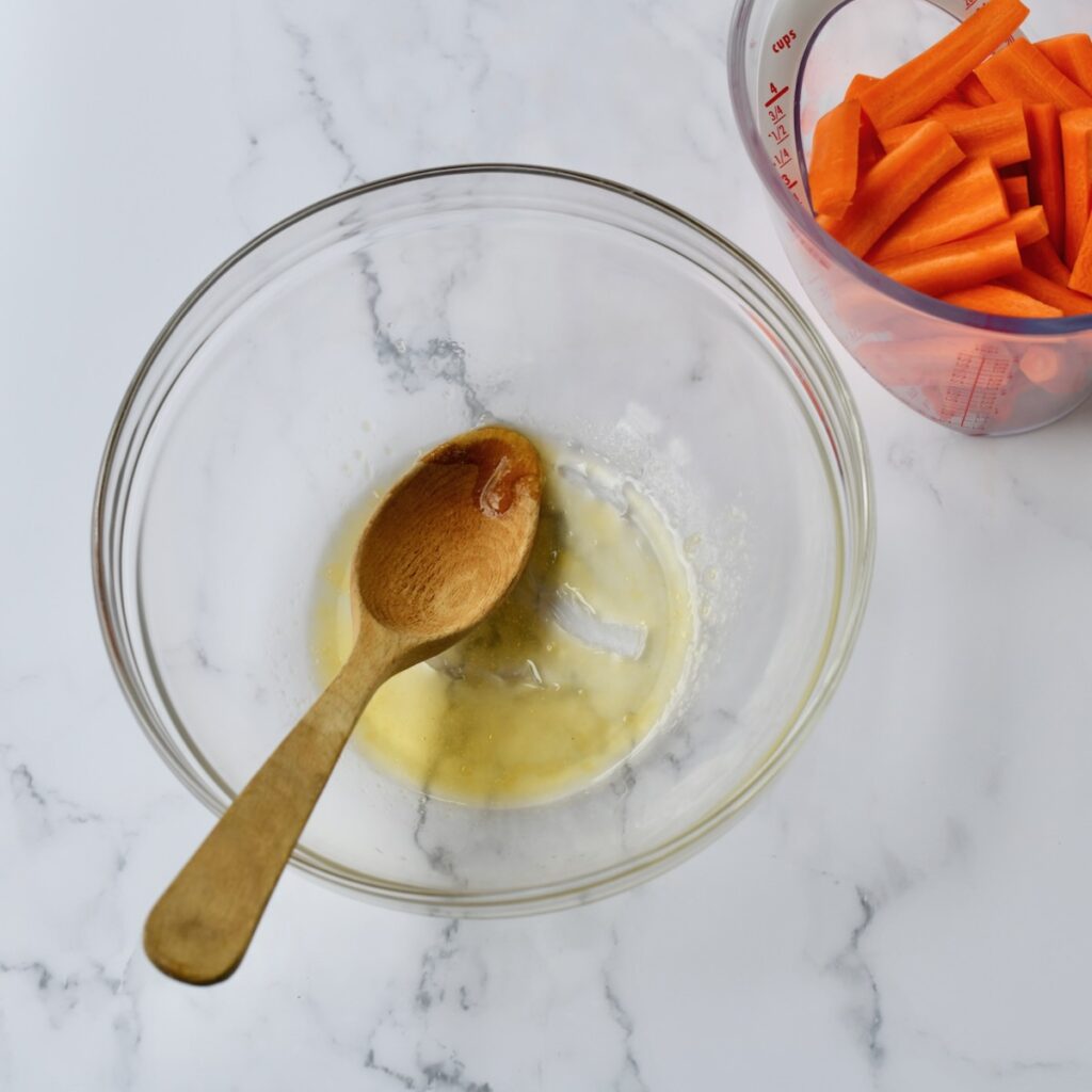 A glass bowl with a honey, salt and oil mixture being stirred with a spoon, with chopped carrots in a measuring cup nearby