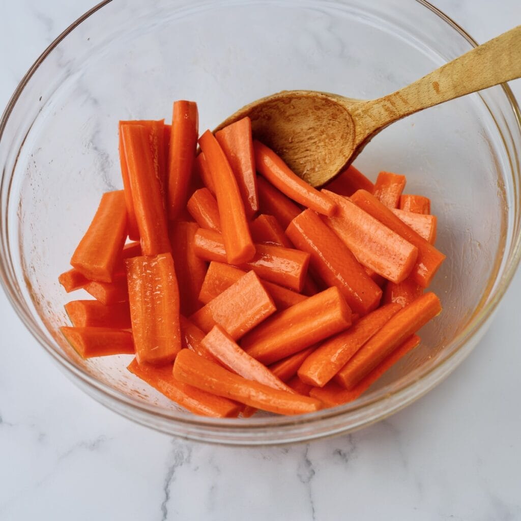 Chopped carrots tossed in a honey, salt and oil mixture inside a glass bowl, stirred with a wooden spoon