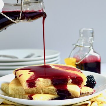 Pouring blackberry syrup over a stack of pancakes on a white plate