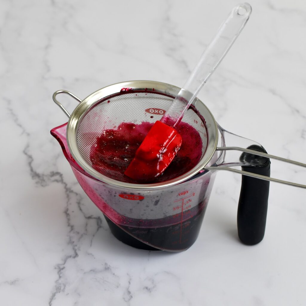 Straining cooked blackberries through a fine mesh sieve into a measuring cup