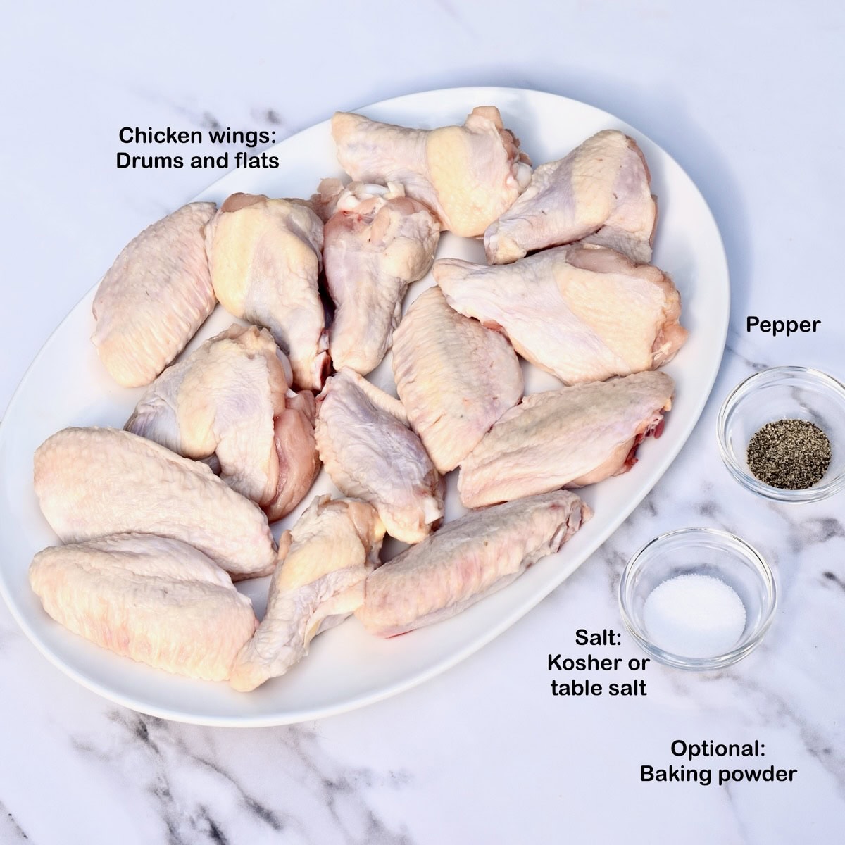 Raw chicken wings on a white plate with small bowls of salt, pepper, and baking powder.