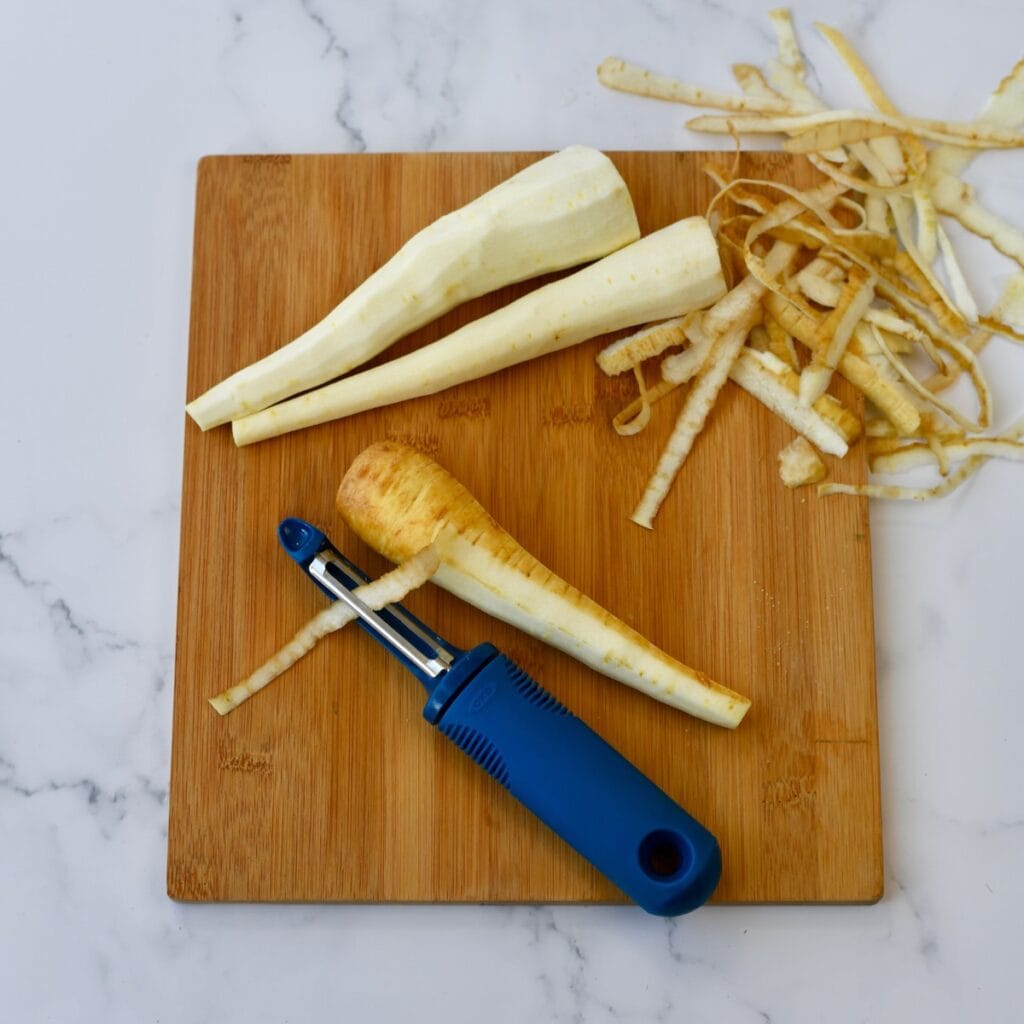 Peeled parsnips and a vegetable peeler on a wooden cutting board