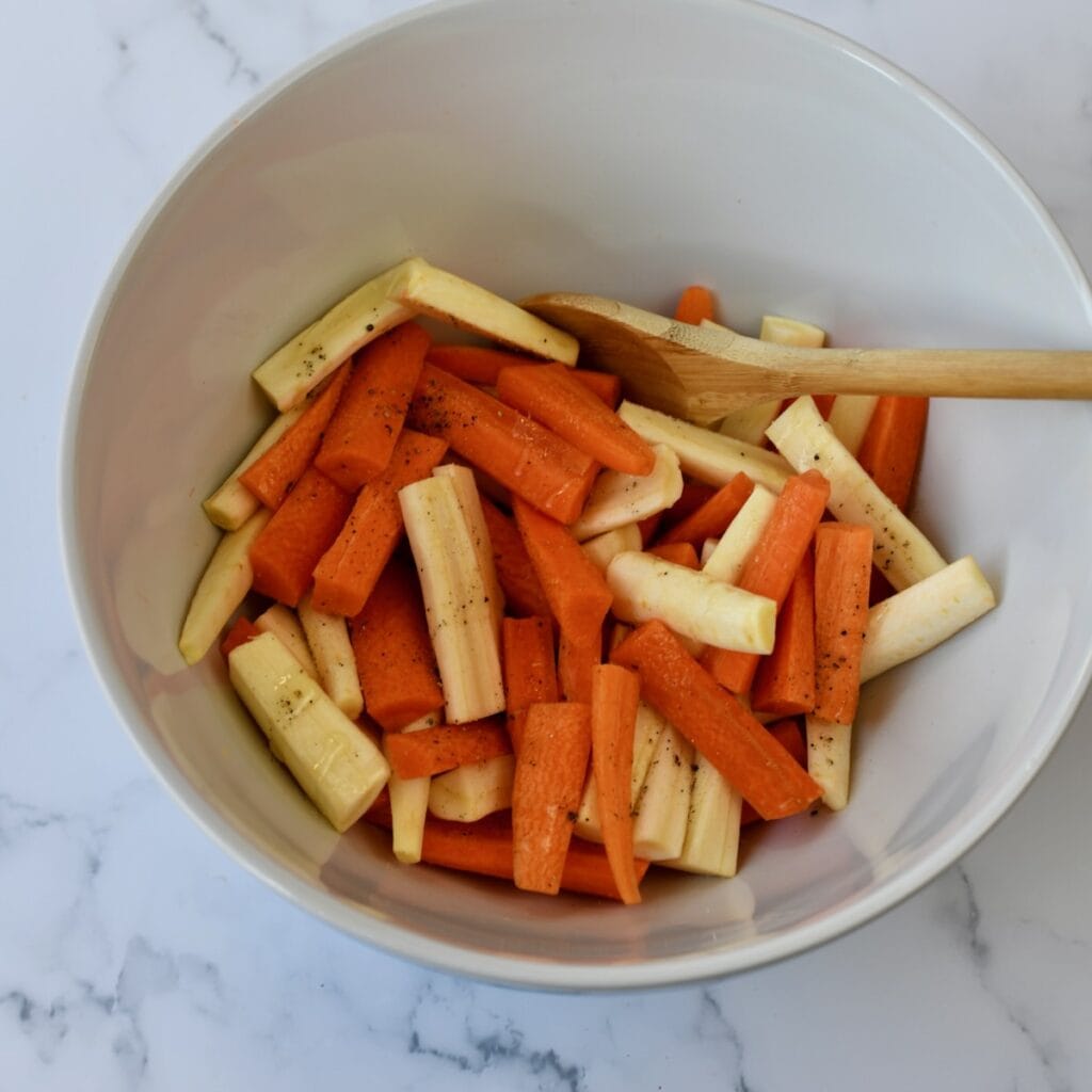 Chopped carrots and parsnips tossed in seasoning in a white mixing bowl