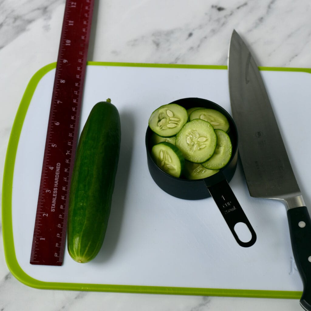 whole Cucumber on cutting board and sliced cucumbers in a measuring cup.