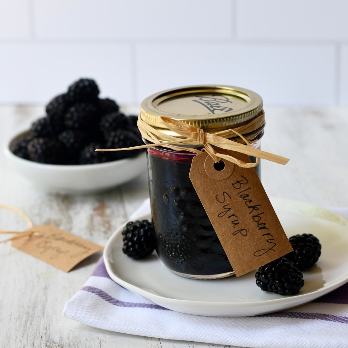Half pint gift jar of homemade blackberry syrup on a plate with fresh blackberries.
