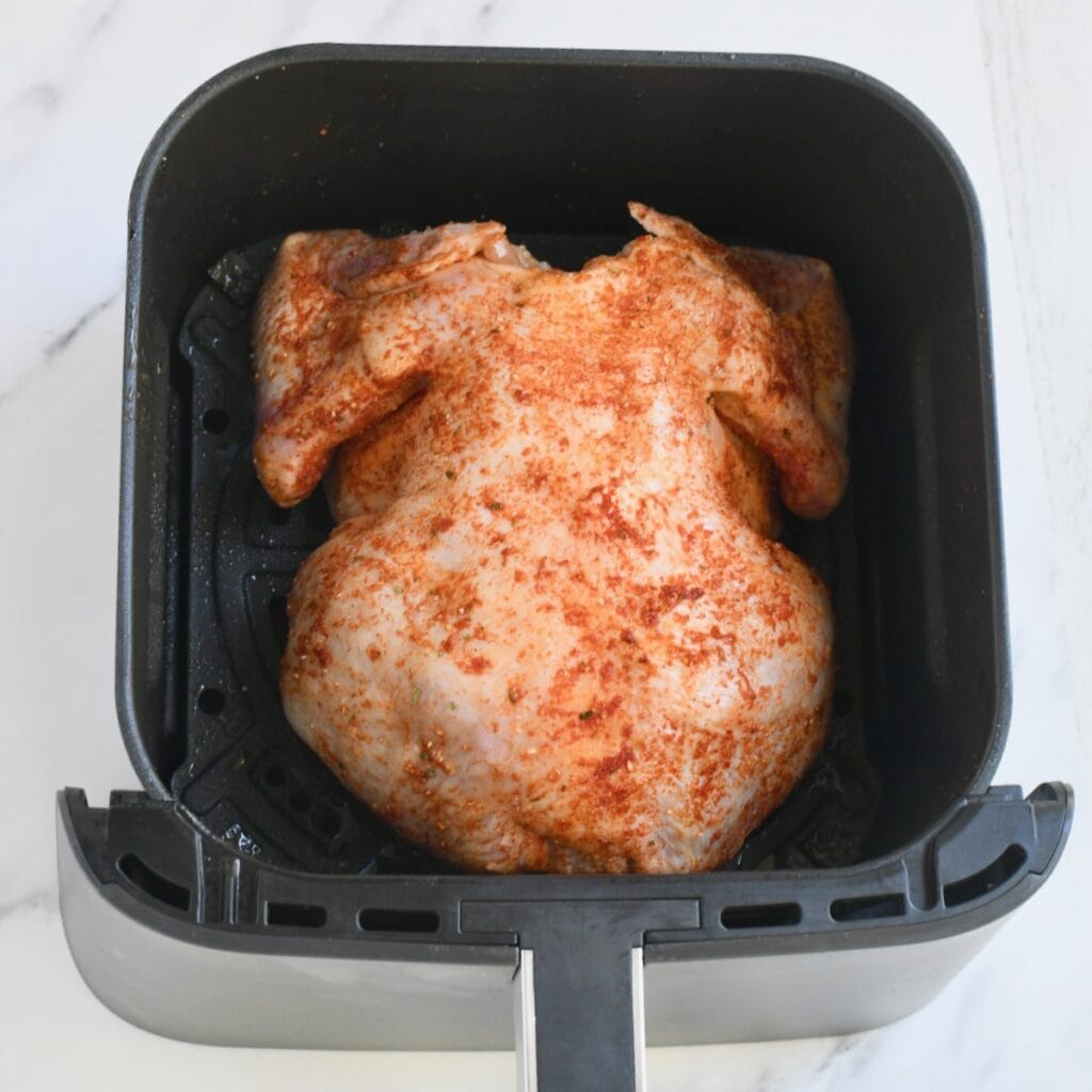 Seasoned raw whole chicken placed breast side down in a Ninja air fryer basket. 
