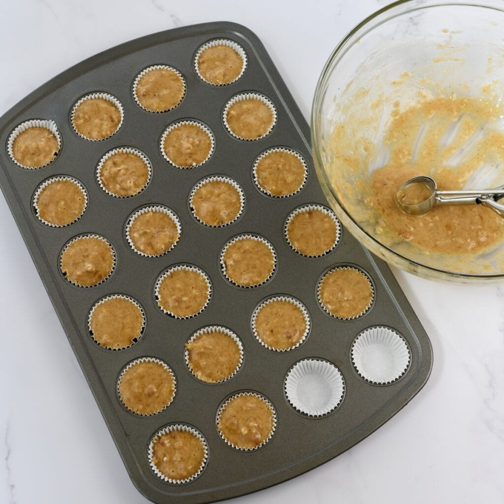 Banana bread batter in mini muffin pans with a cookie scoop in the batter bowl.