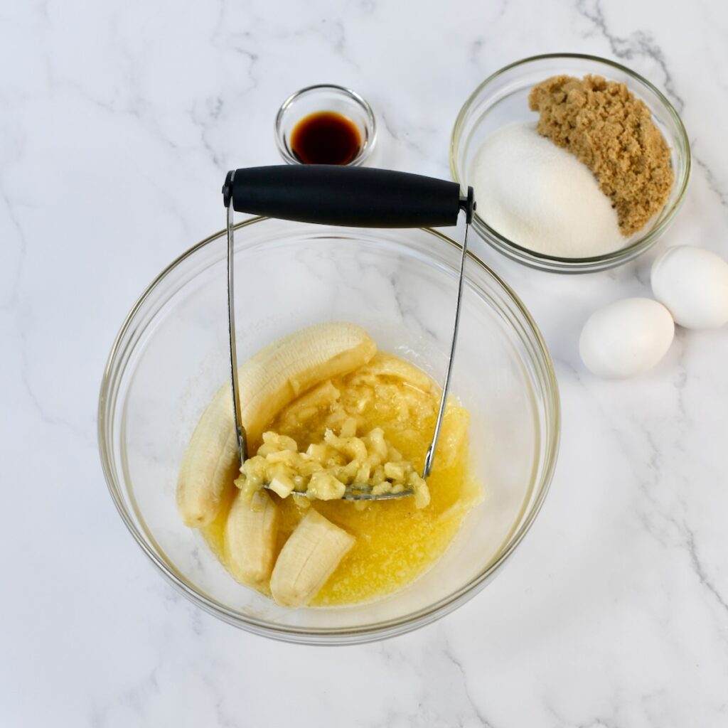 Oxo potato masher mashing ripe bananas in a glass bowl.

