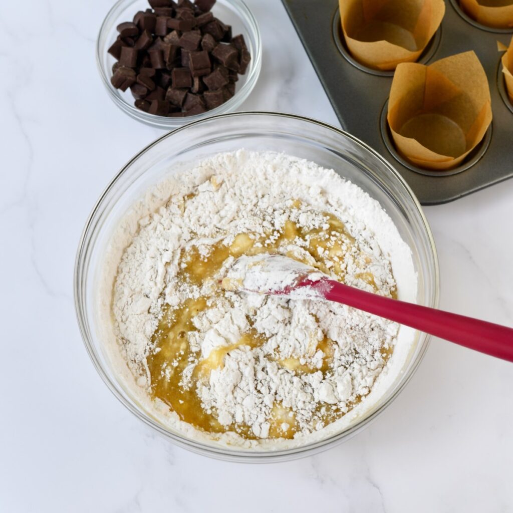 Red spatula blending dry ingredients into mashed banana batter.
