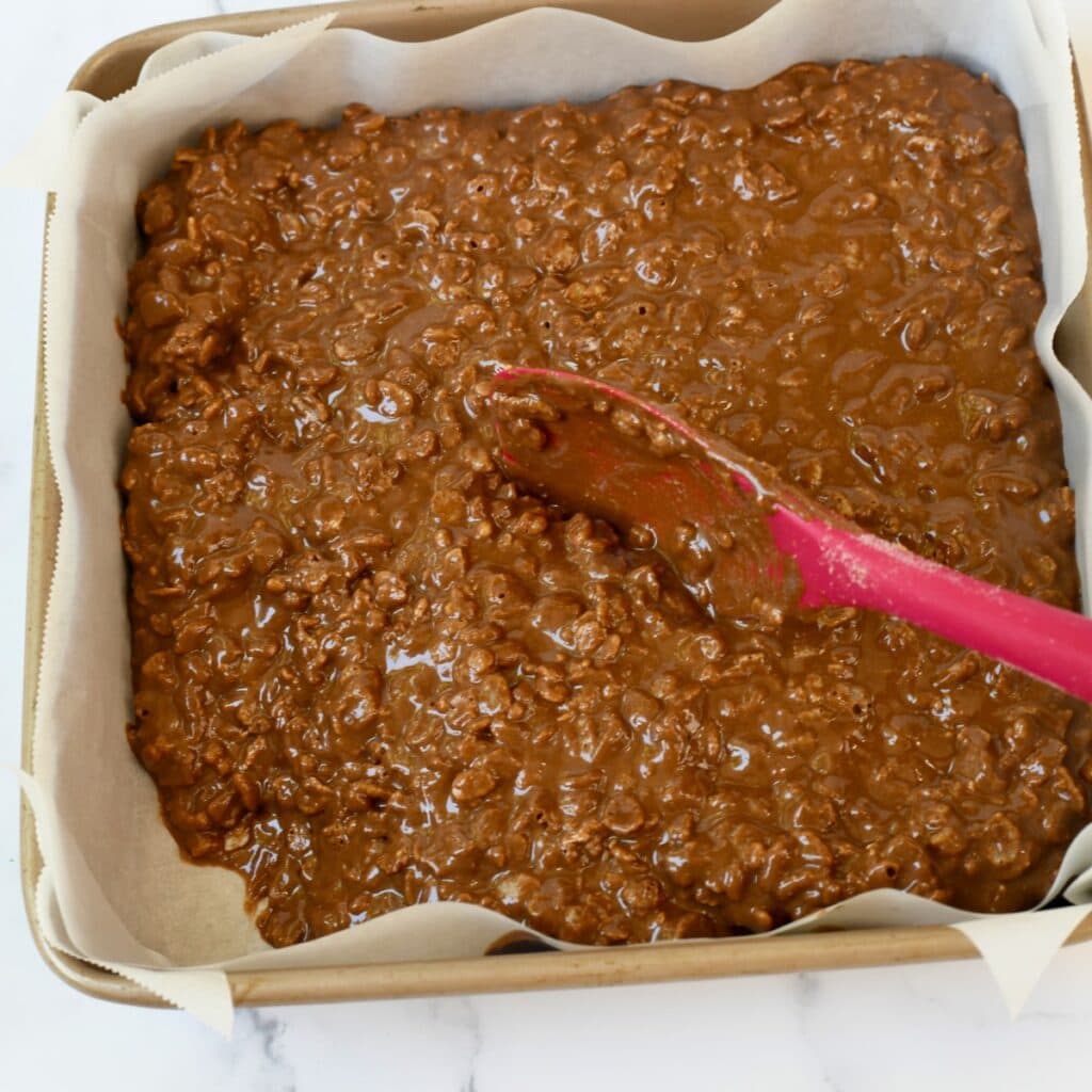 Red spatula spread out chocolate peanut butter rice krispie treats batter in pan.