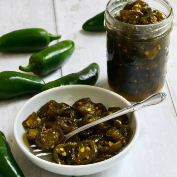 A white bowl of candied jalapenos with tiny fork in it next to a jar of cowboy candy with jalapeno peppers on the surface.