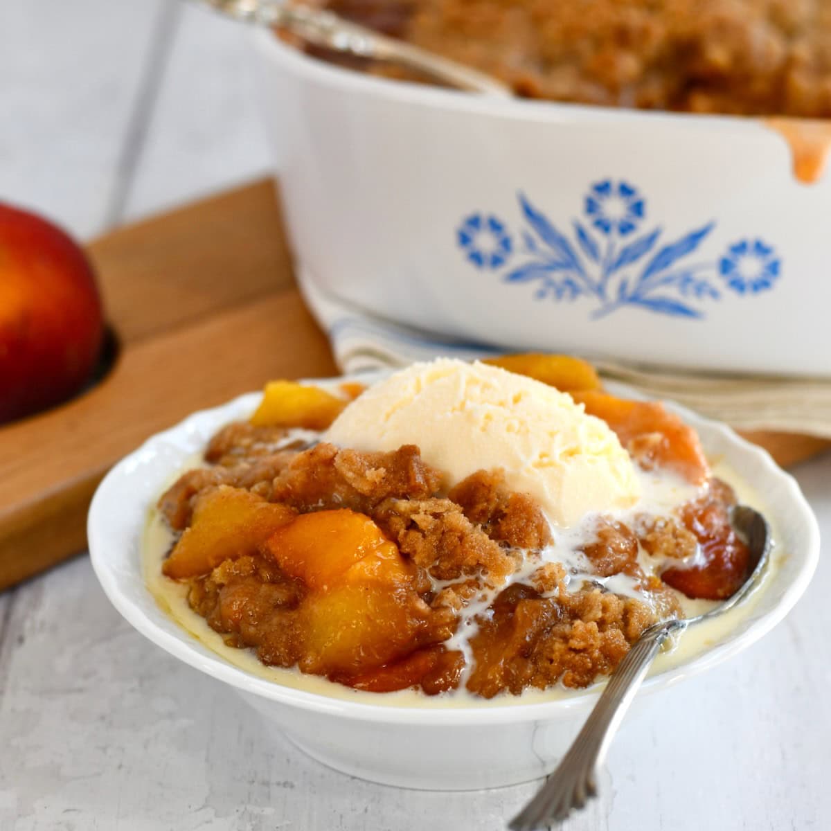 White bowl full of fresh peach crumble topped with melting vanilla ice cream.
