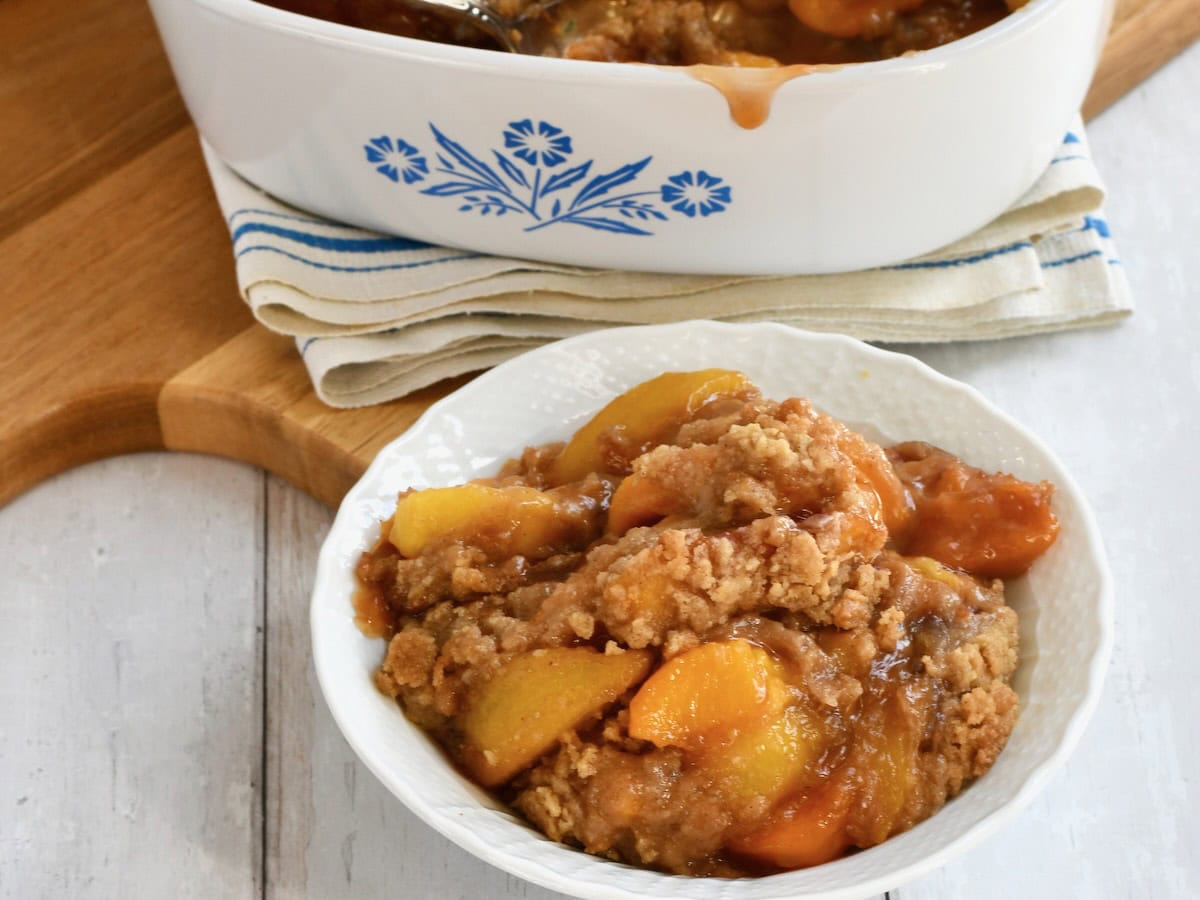 Peach crumble in white bowl with casserole dish behind it. 