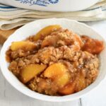 Peach Crumble Cobbler piled high in a white bowl next to blue and white cloth.