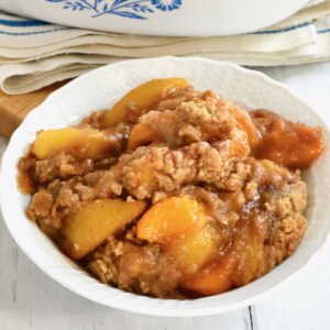 Peach Crumble Cobbler piled high in a white bowl next to blue and white cloth.