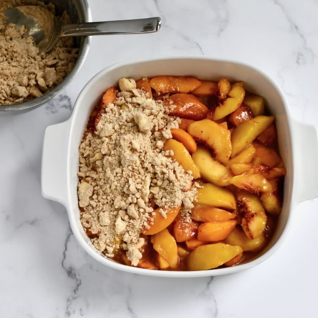 White Pyrex casserole dish with peach cobbler mixture topped with unbaked crumble topping. 