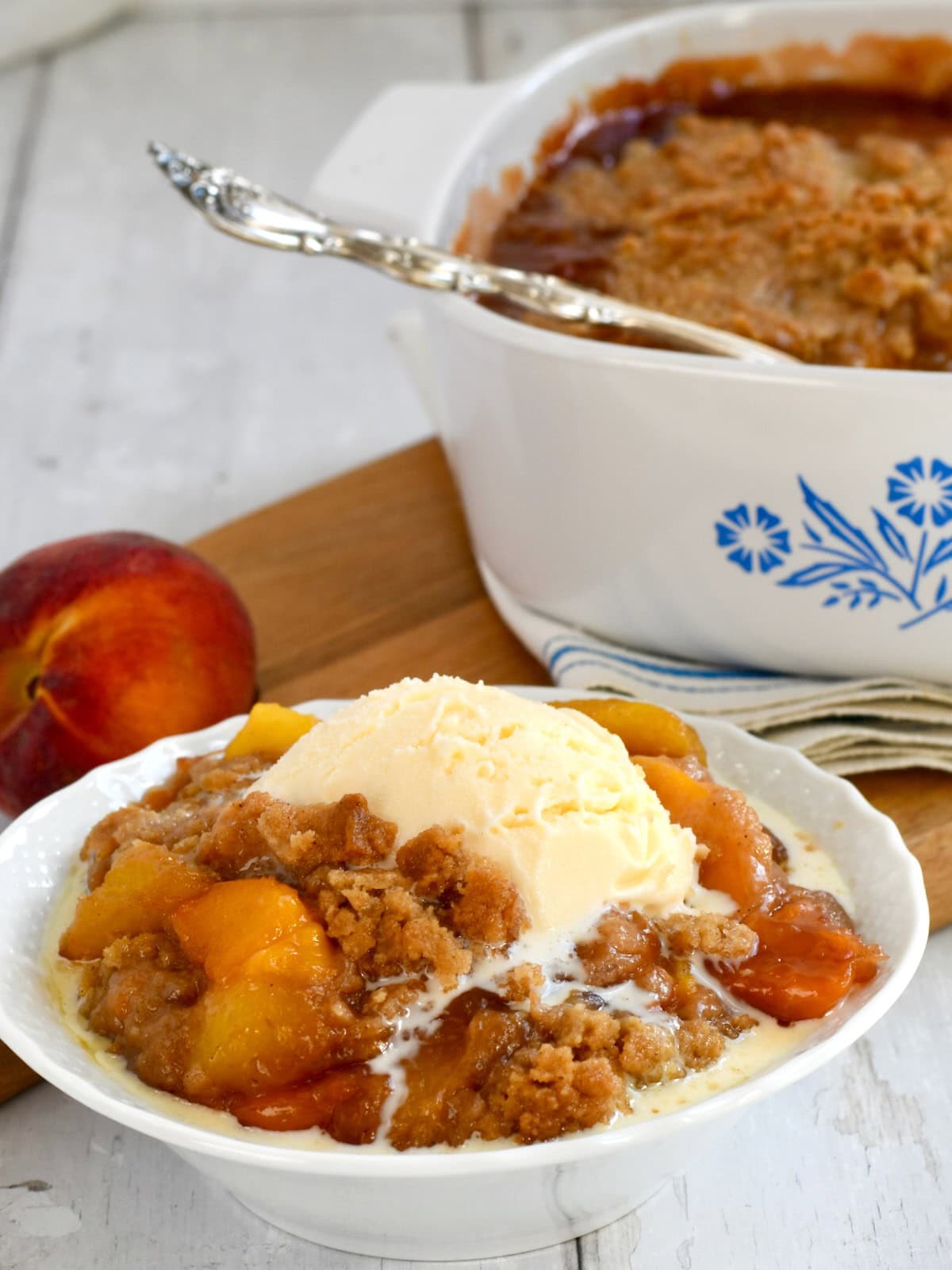 Fresh peach crumble with vanilla ice cream and casserole dish in background.
