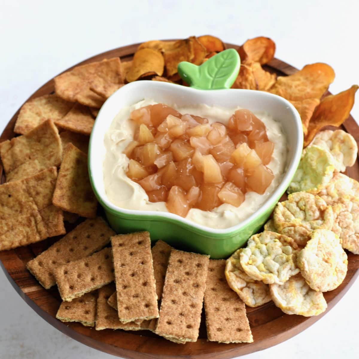 Apple pie dip in a green apple-shaped bowl topped with chopped apple pie filling, served on a wooden platter with graham crackers, rice cakes, and assorted chips