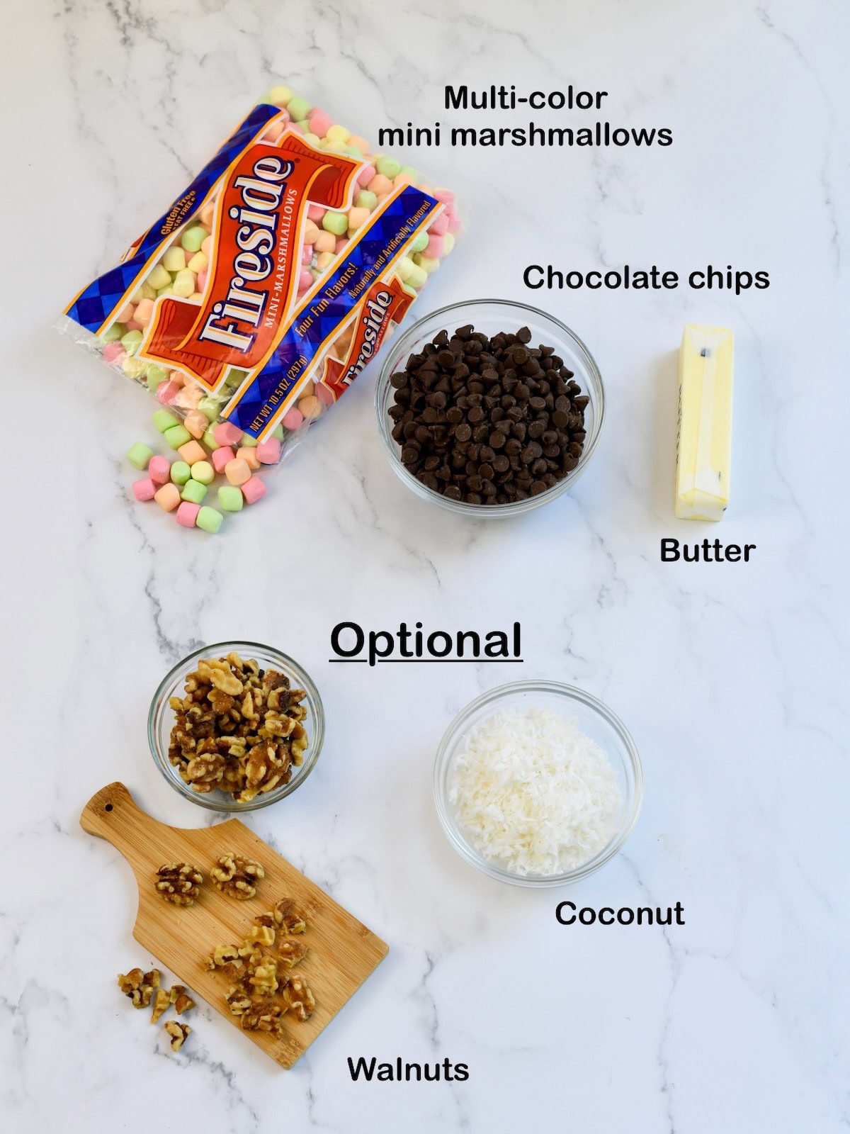 ingredients for cathedral windows recipe on a marble surface including mini marshmallows, chocolate chips, butter, coconut, and walnuts