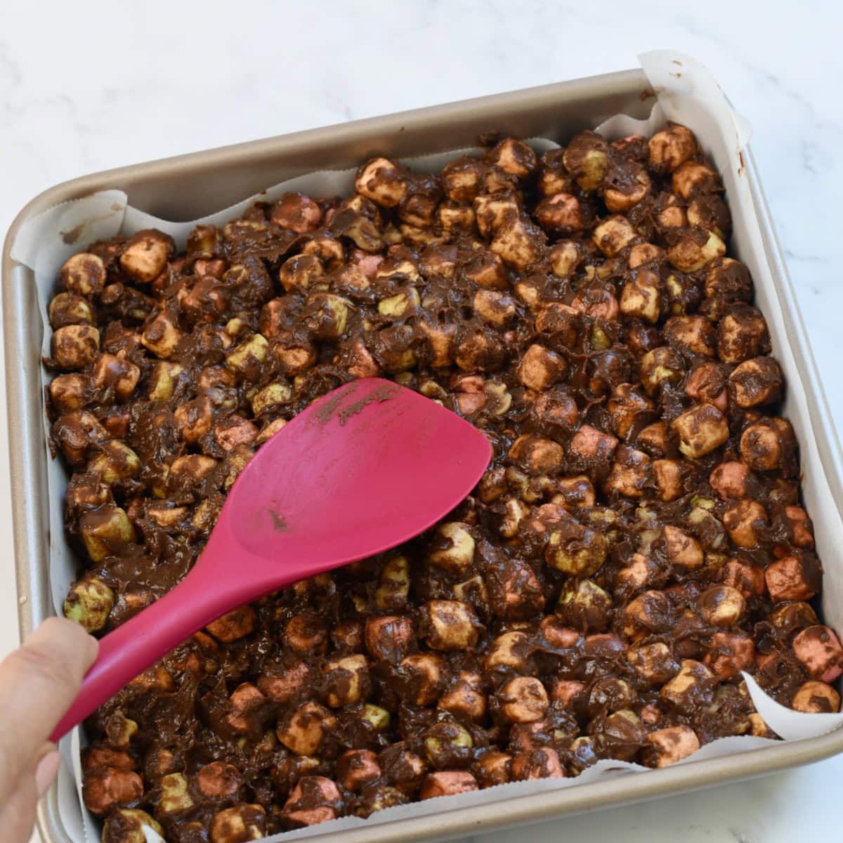 spreading chocolate and marshmallow mixture in a square baking pan lined with parchment paper using a red spatula