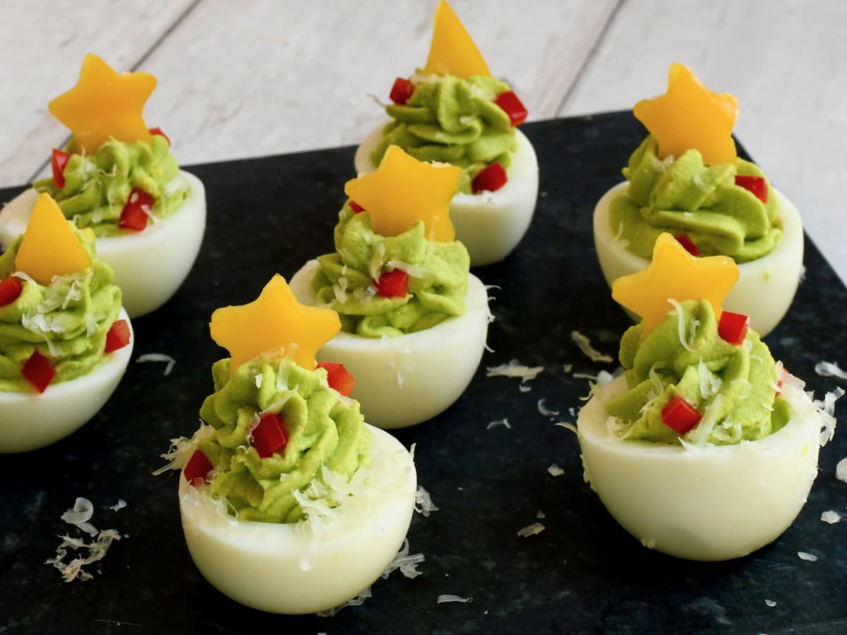 christmas tree deviled eggs topped with yellow bell pepper stars, red pepper ornaments, and a sprinkle of parmesan snow on a black serving board